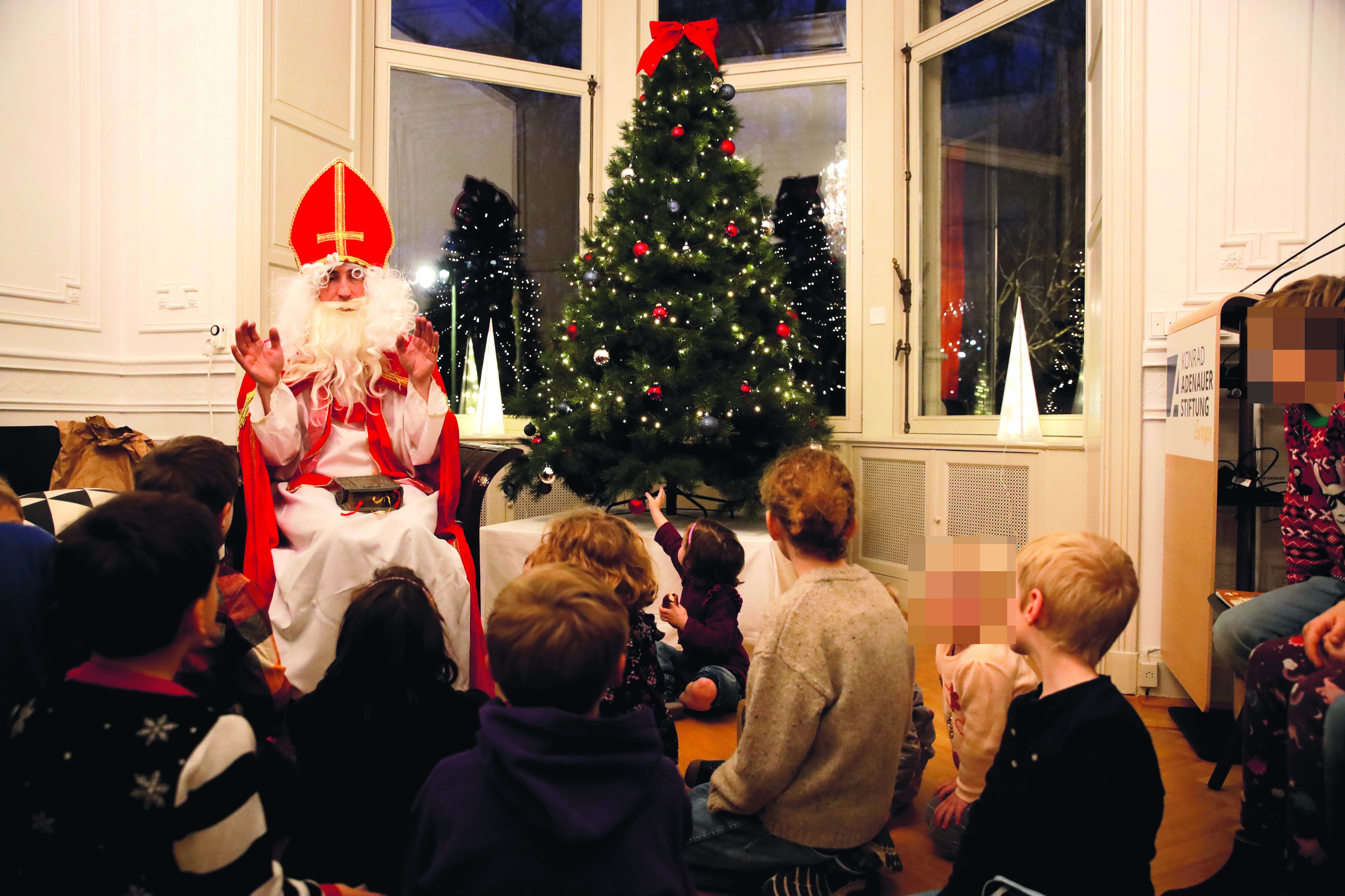 Nikolaus sitzt in vollem Ornat vor einem Weihnachtsbaum und erzählt mit gehobenen Händen eine Geschichte. Um ihn herum sitzen 20 Kinder (davon 13 auf dem Photo - Gesichter verpixelt) und lauschen.
